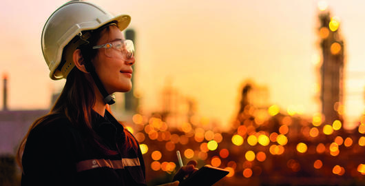 A woman in a safety hat makes notes on a tablet against a cityscape backdrop, showing commitment to safety