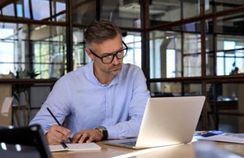 man sitting in front of open laptop writing on a pad while in an office